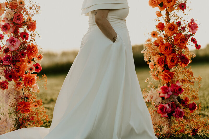 Mariée de profil, mains dans les poches dans une prairie entourée d'une arche de fleurs rouges, oranges et roses. Prise par Laura Termeau Photographie.