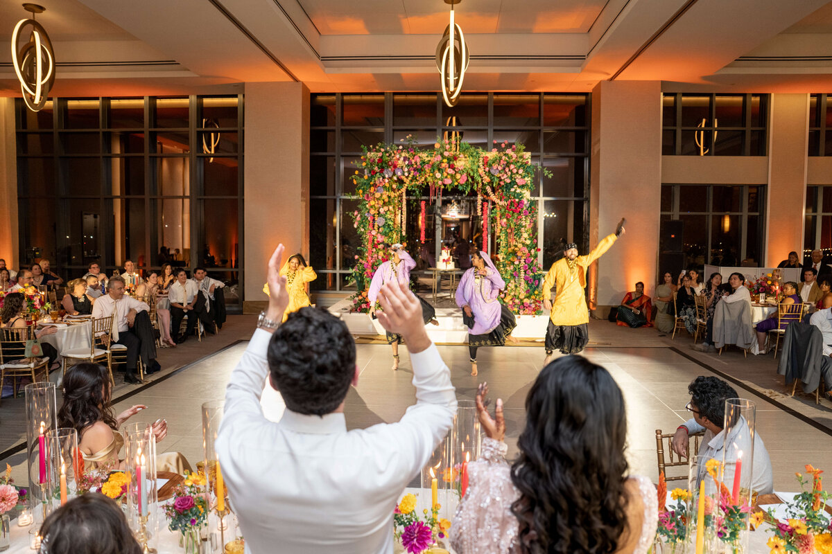 A group of people in traditional attire perform a dance on a well-lit stage at a vibrant indoor celebration, while an audience seated at decorated tables watches. The scene is festive, with colorful flowers and warm lighting throughout the venue.