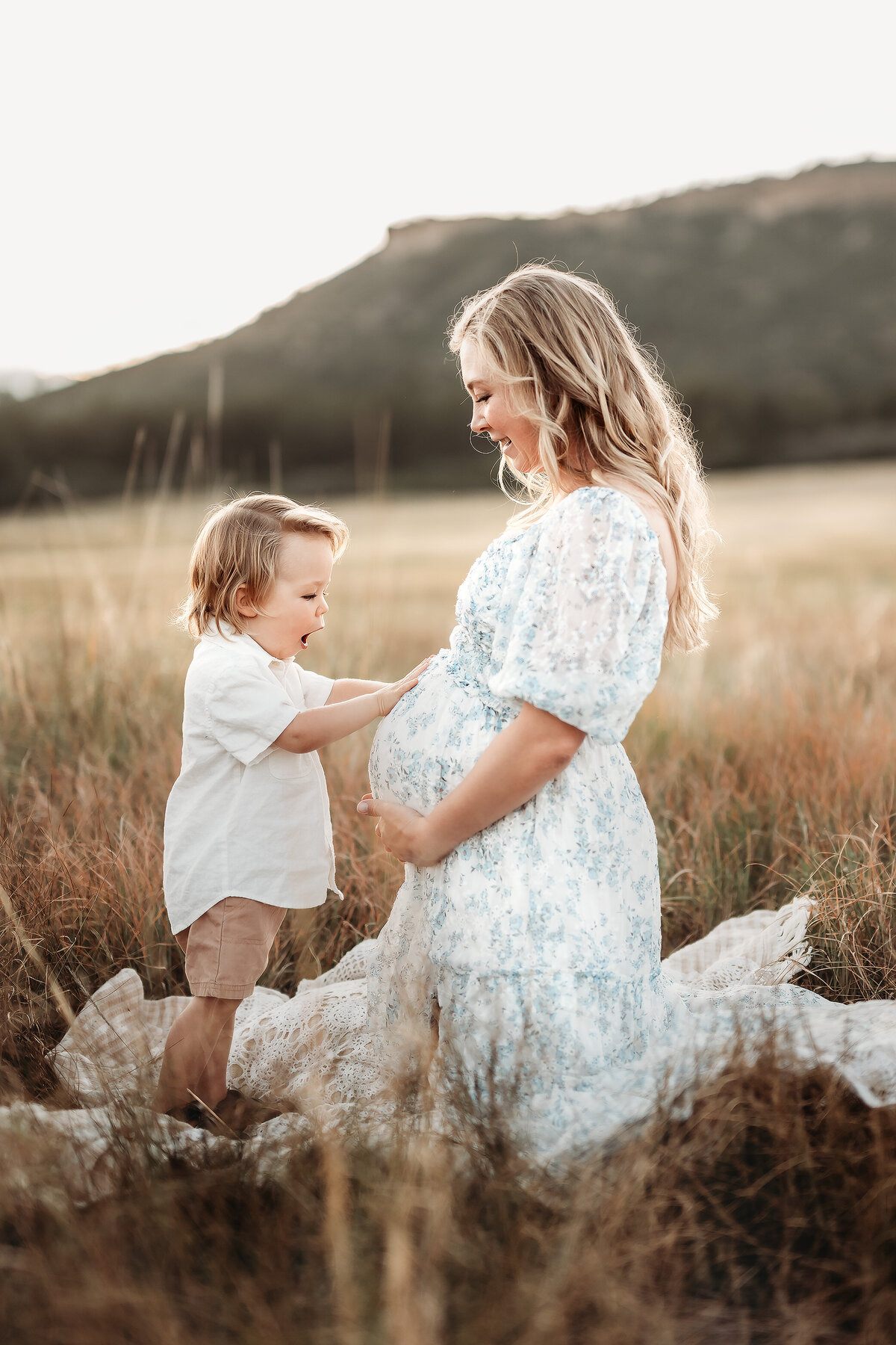 pregnant mother kneeling in field with son touching her pregnant belly with a surprised face