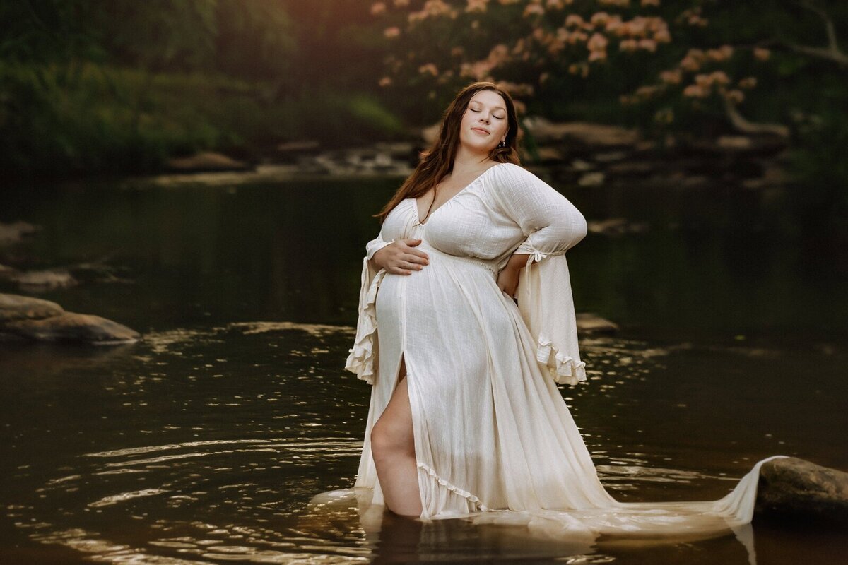 Pregnant woman in a flowing white dress stands in a shallow river, surrounded by greenery and softly lit by sunlight.