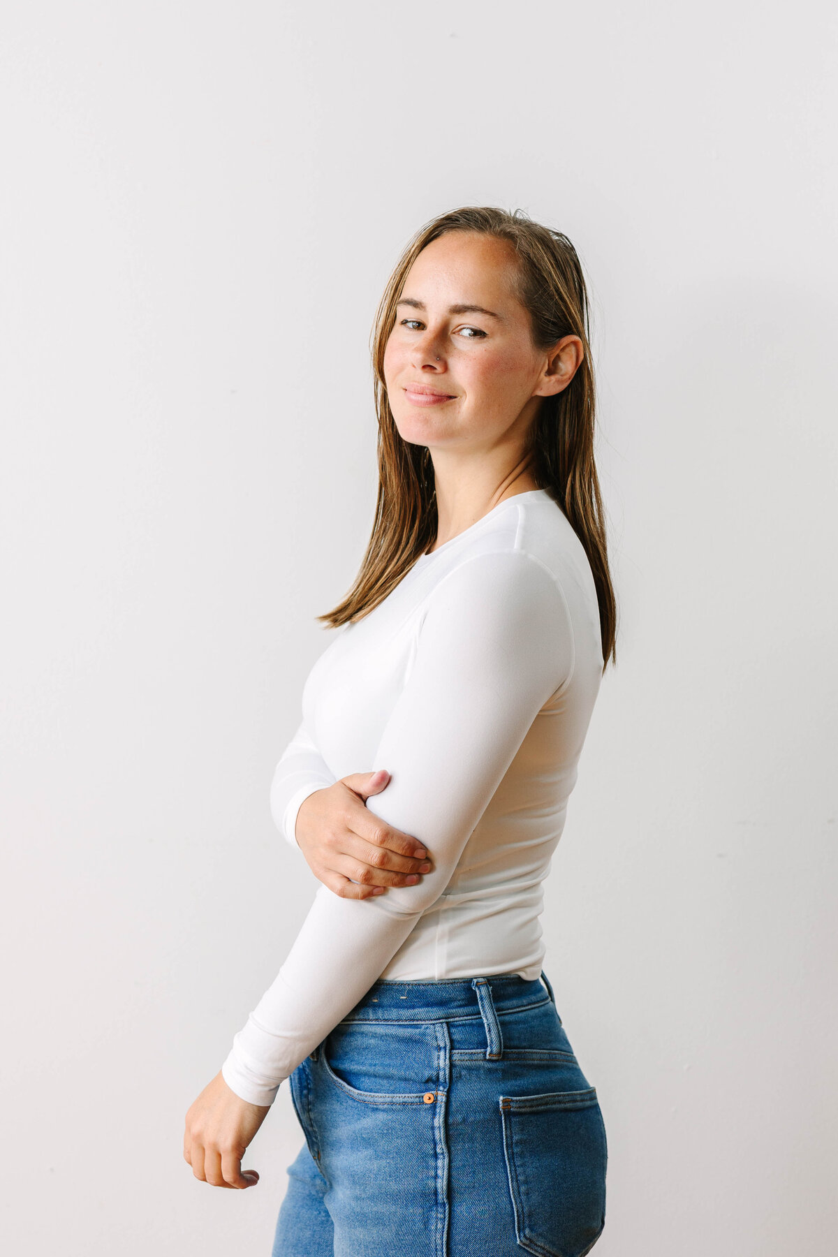 A woman with long brown hair poses against a plain white background, wearing a fitted white long-sleeve top and blue jeans. She looks slightly over her shoulder with a soft smile, her arms crossed casually.