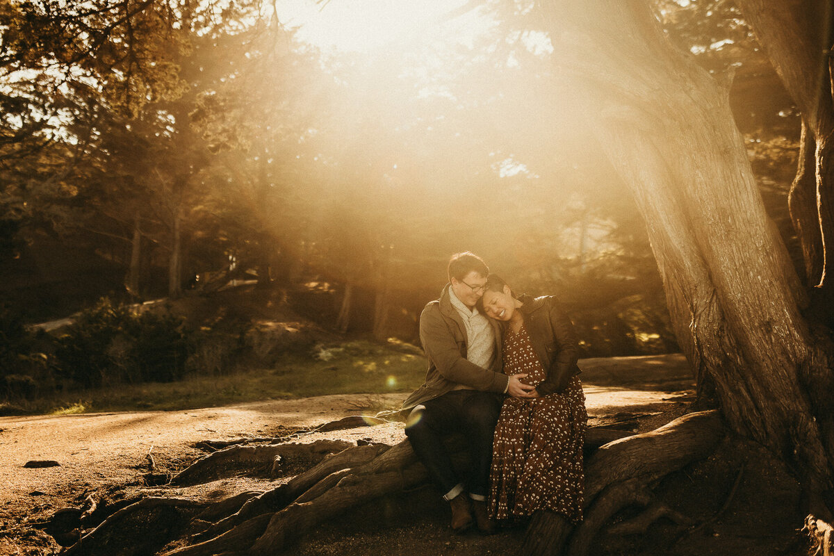 Bay area maternity couple in wooded area with bright sunbeams of light and snuggling pregnant belly while sitting side by side