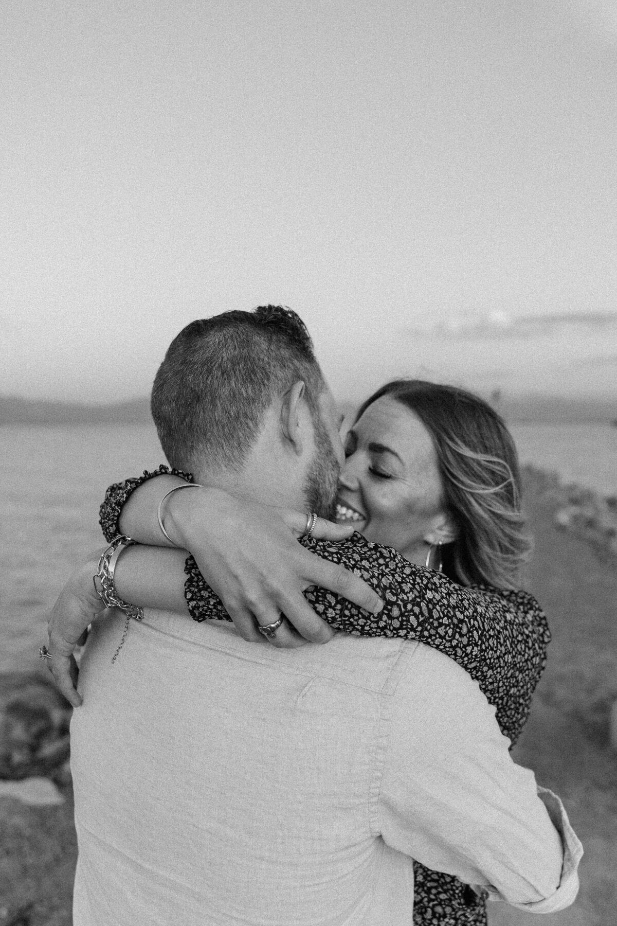 A black and white image of a couple embracing near Utah Lake, their faces full of joy and love as they hold each other closely.