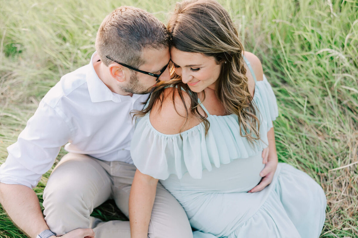 husband and wife touching foreheads and cuddling in a field. She is leaning back on him and she has her hand on her belly and is wearing a green dress.