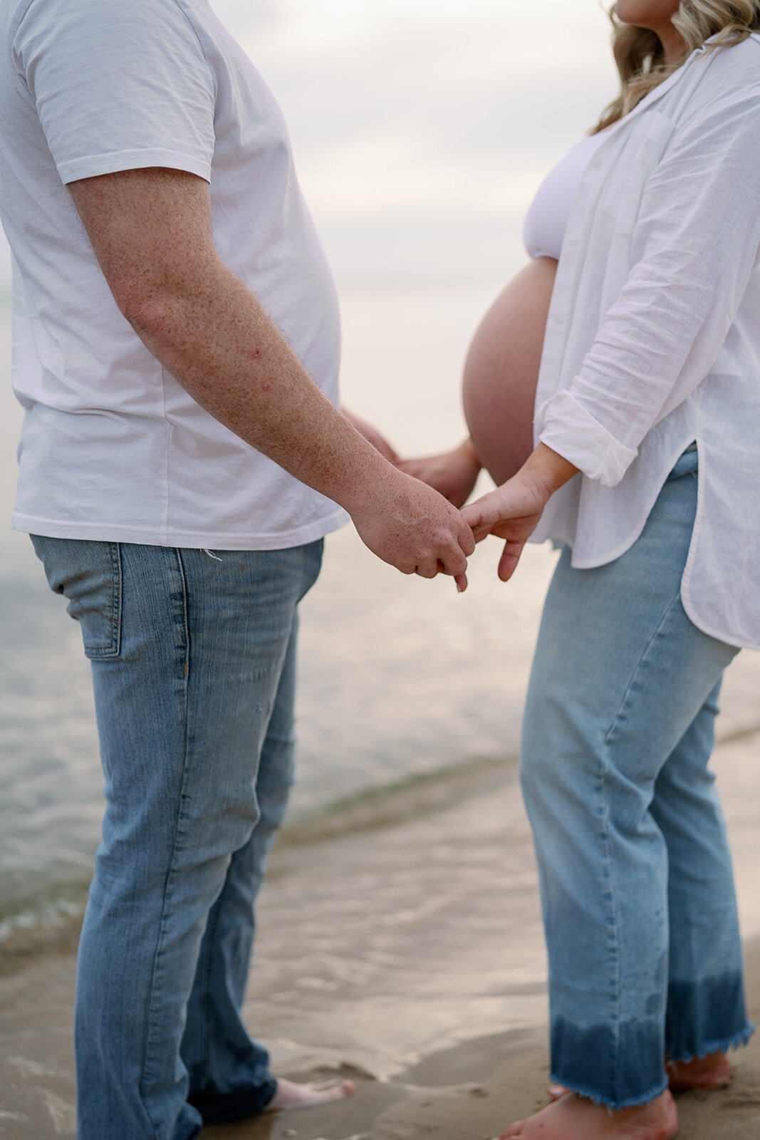 Expecting couple holding hands by the water’s edge during maternity photoshoot at South Haven North Beach in Michigan.