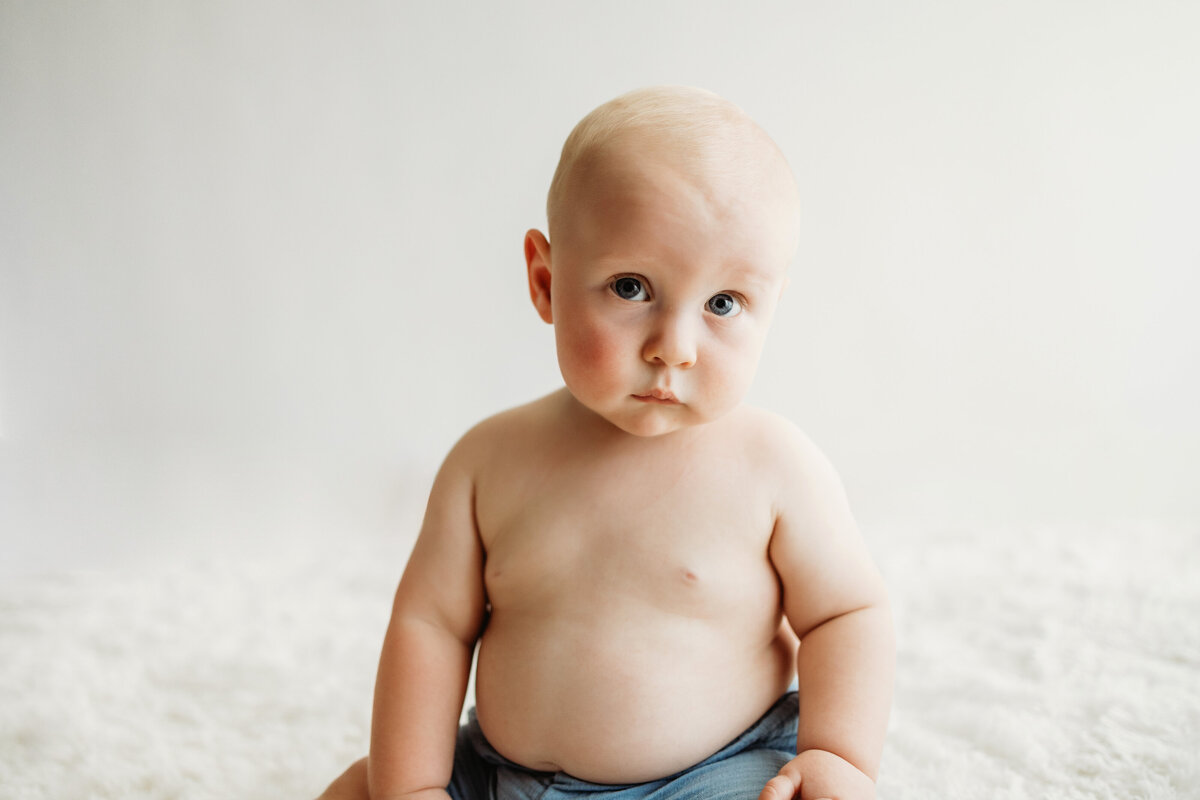 An infant six month old baby boy sitting in just a diaper with a white background for his sitter milestone session