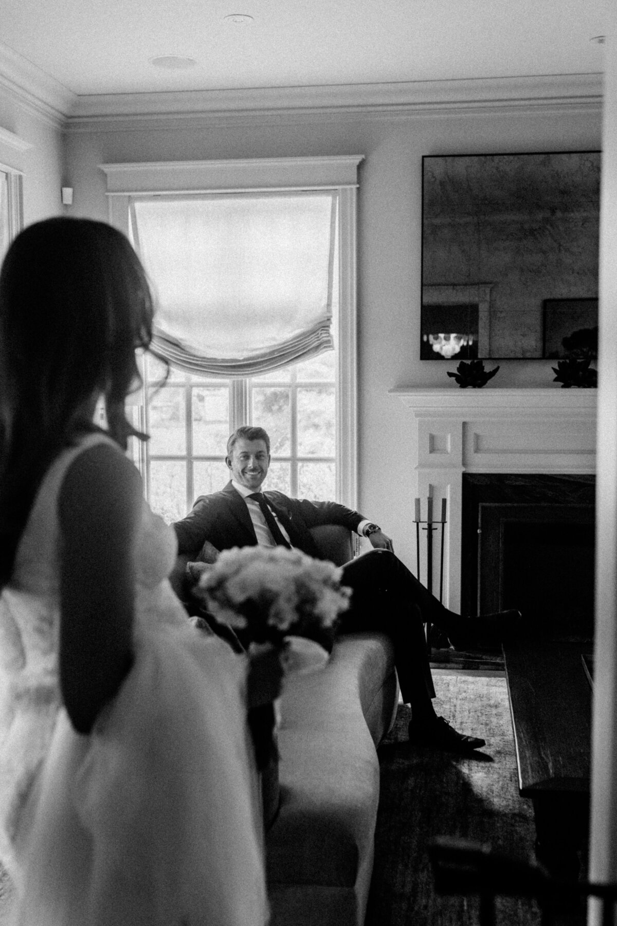 A sophisticated black-and-white image of a pregnant bride approaching her groom, who is seated and smiling, capturing the anticipation of their first look