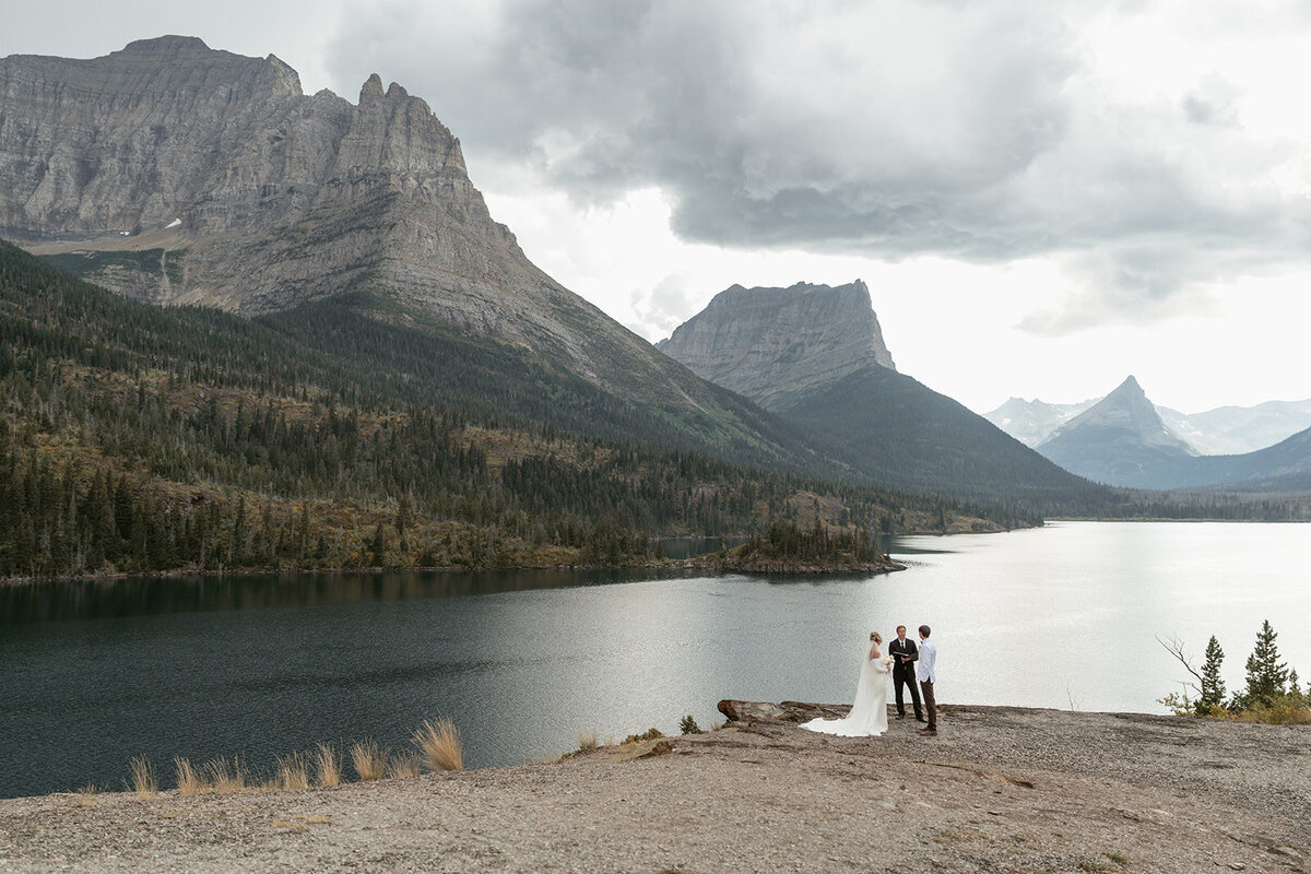 A couple stands together on a rocky overlook above a glacier-fed lake surrounded by dramatic mountain peaks and moody skies in Glacier National Park, captured by Sydney Breann Photography during their intimate Montana elopement.