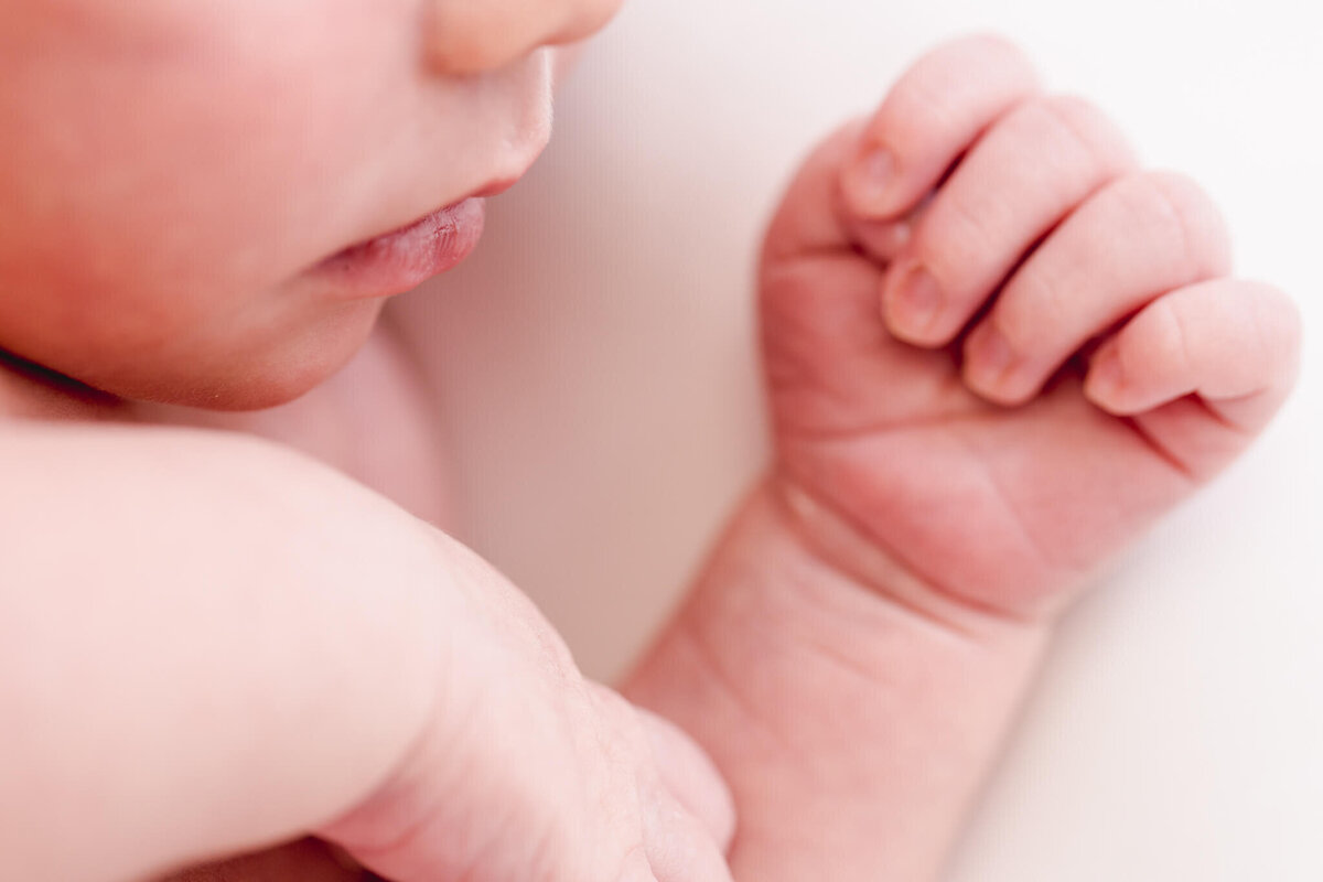 Close-up of a babys face and hand. The image focuses on the softness of the skin and the gentle curve of the lips, with the fingers curled slightly, conveying a sense of tenderness and innocence.