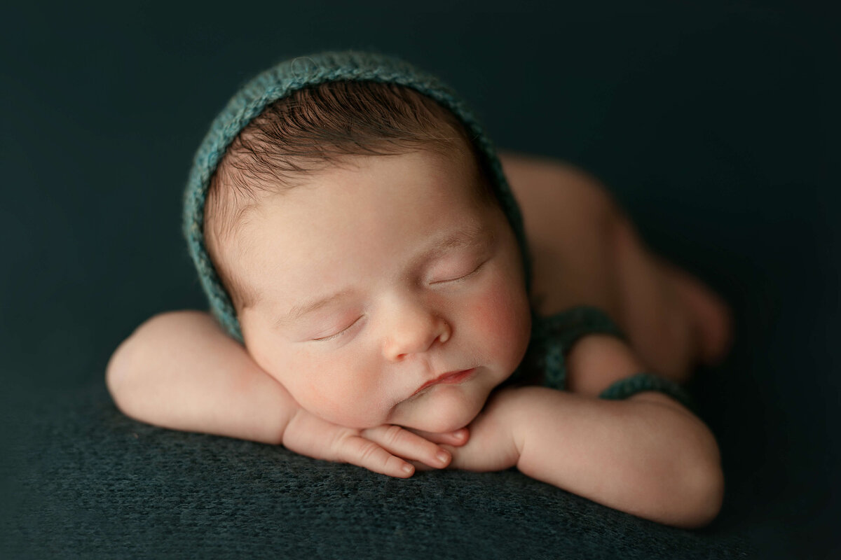 newborn boy in a teal bonnet laying with his hands on his head at a newborn photo session