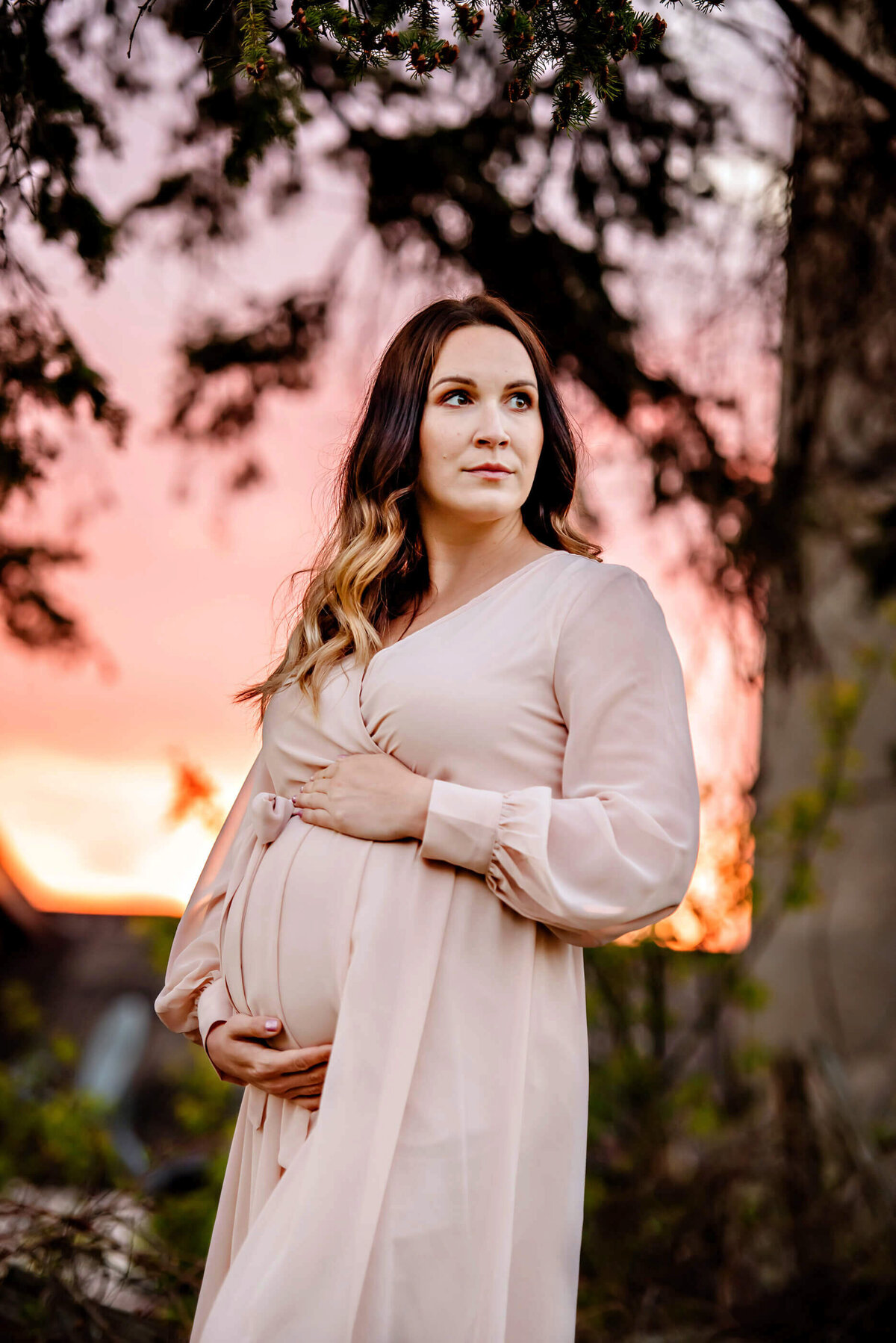 pregnant woman wearing a pink dress with a pink sky behind her for her outdoor maternity photos in Burlington
