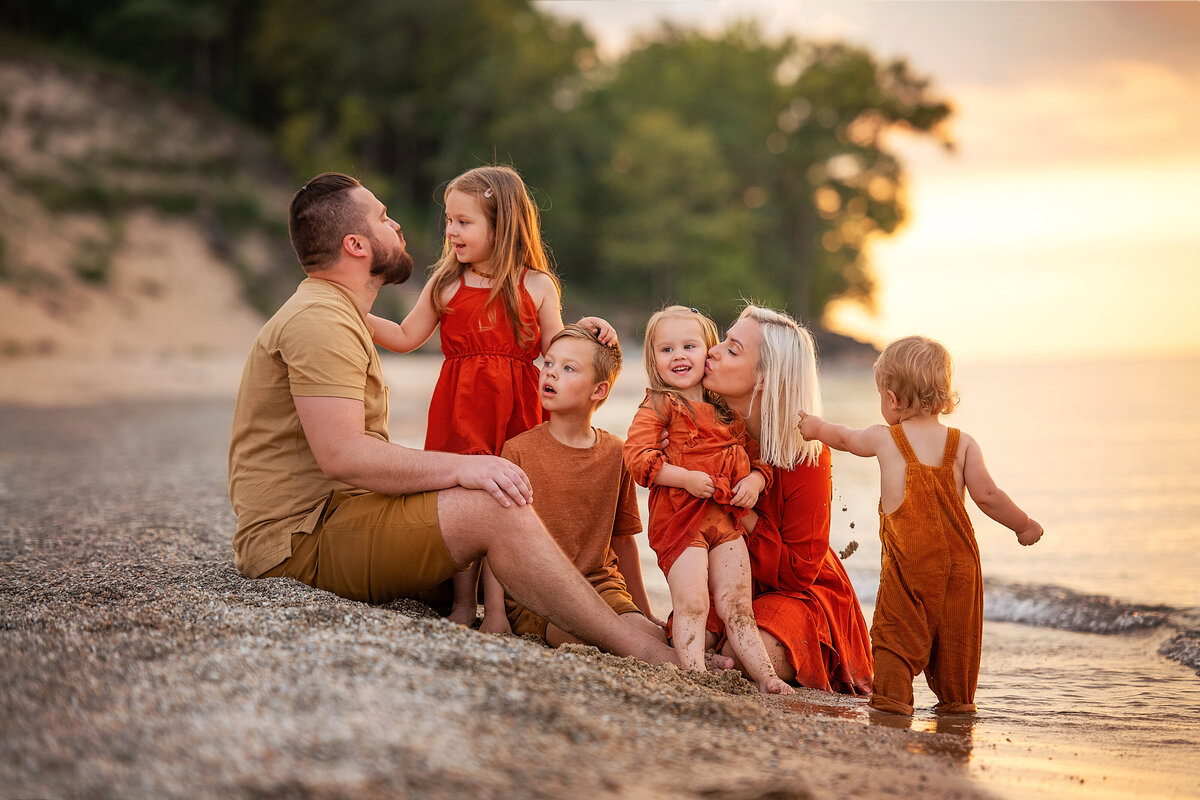 Young parents with four little kids in matching orange outfits sitting on the sand during sunset at the beach.