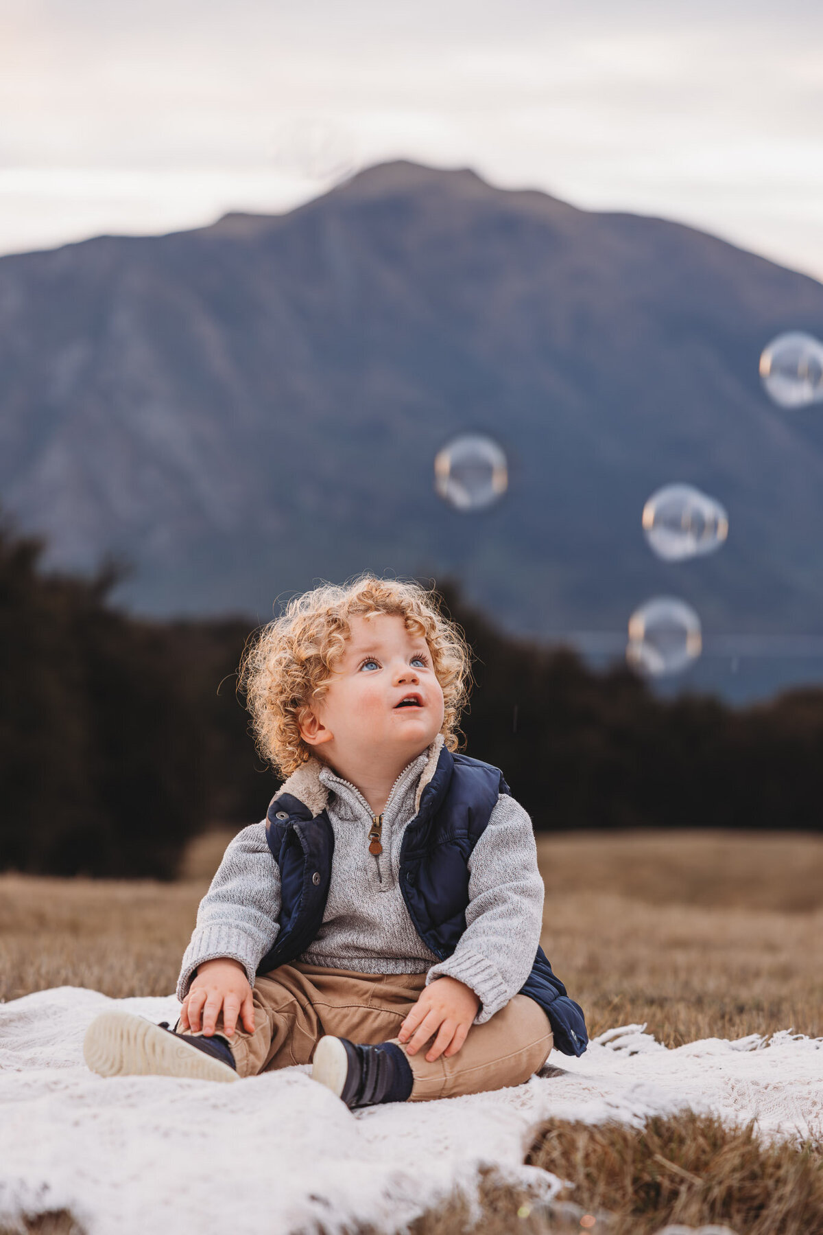 a boy looking at bubbles in the air while sitting on a blanket during a children photography session in wanaka captured by Bie