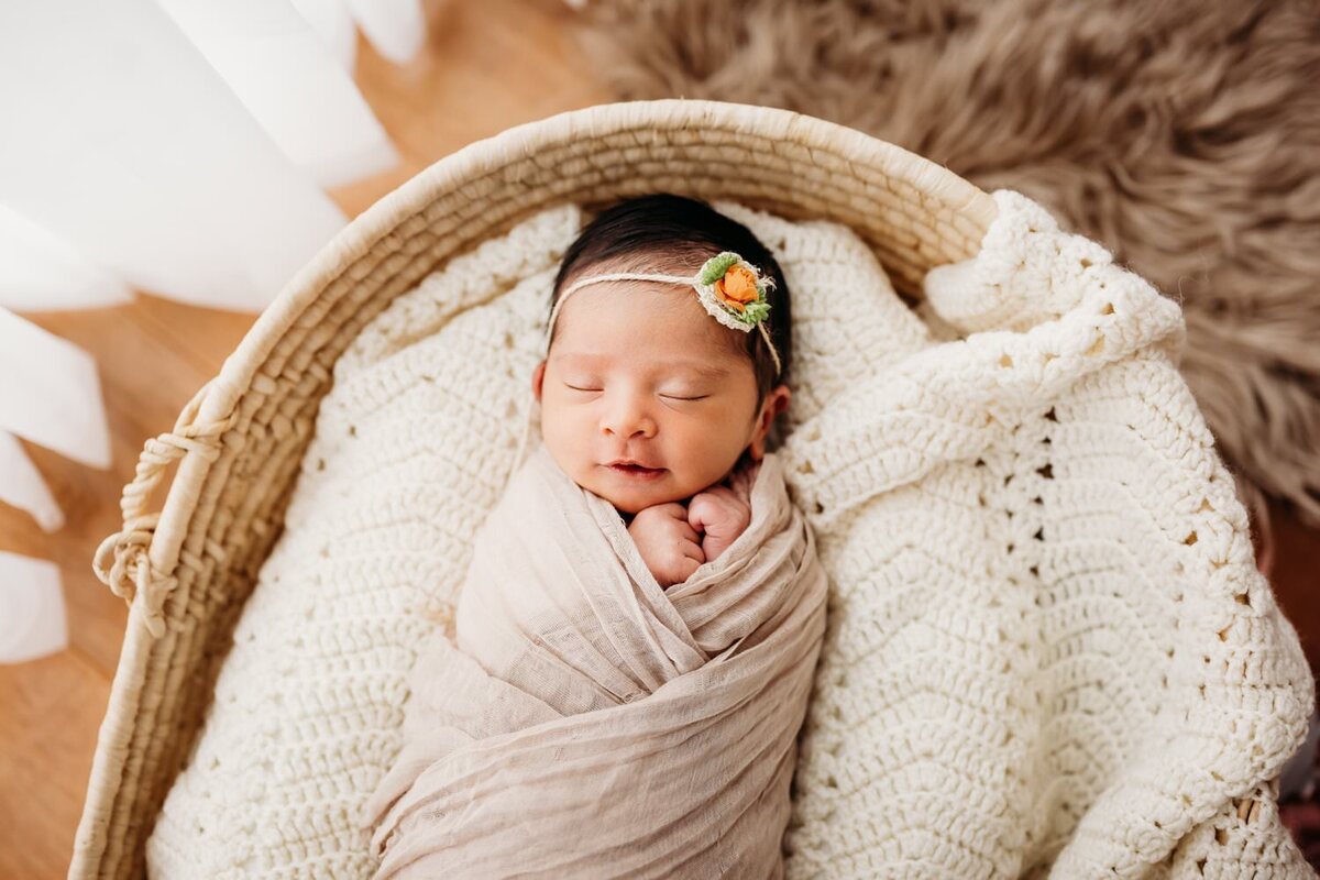 studio photo of baby girls sleeping in moses basket. she wears a flower on her head