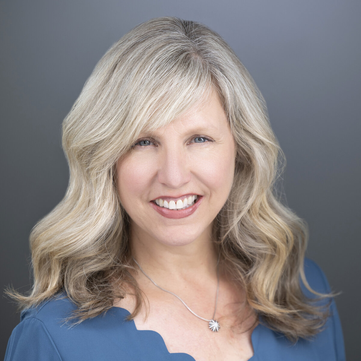 Woman in blue shirt and blond hair smiles at camera with piercing blue eyes for a headshot with a grey background in Sacramento headshot studio.