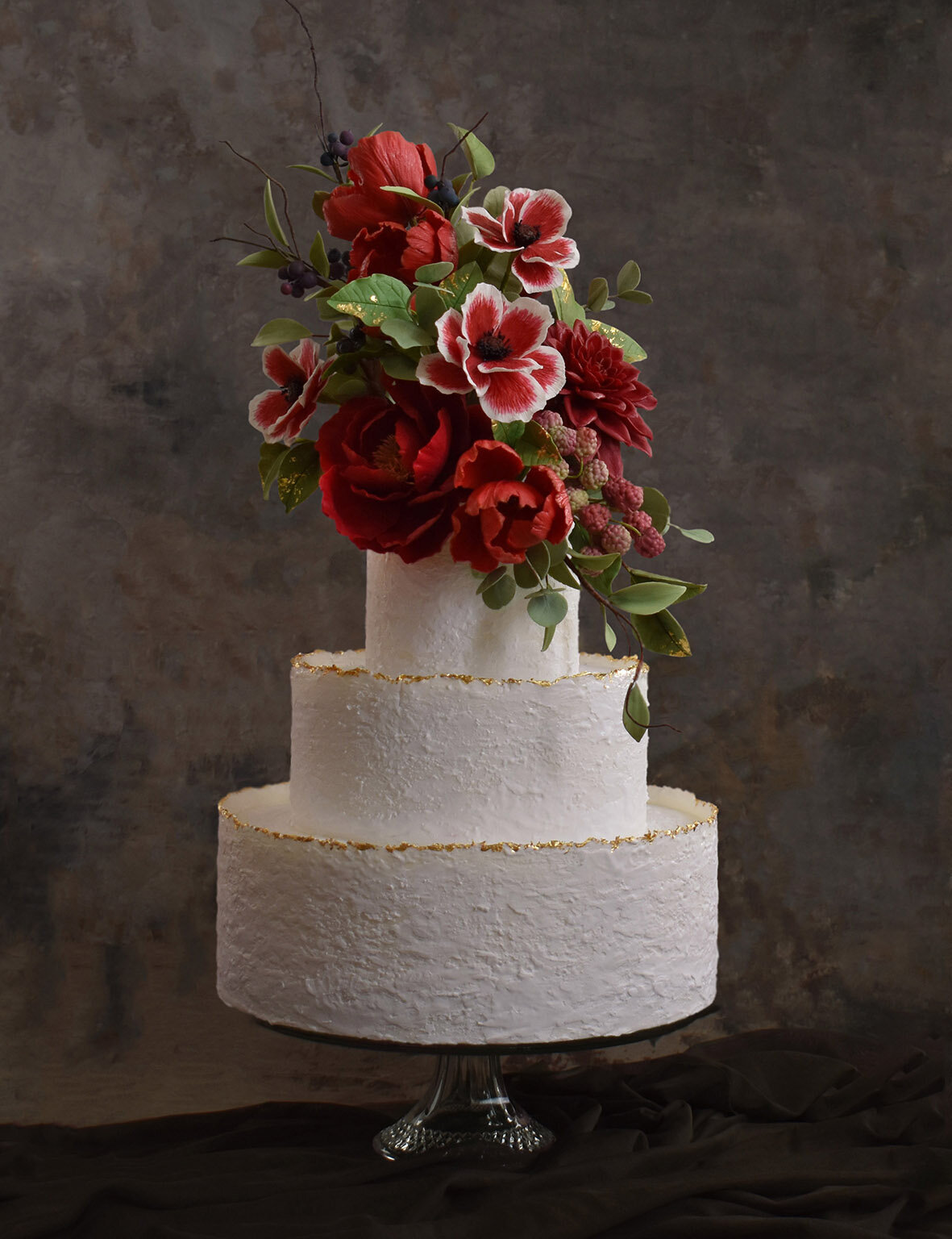 Three tiered white wedding cake with subtle texture, gold leaf detail and a dramatic display of deep red sugar flowers