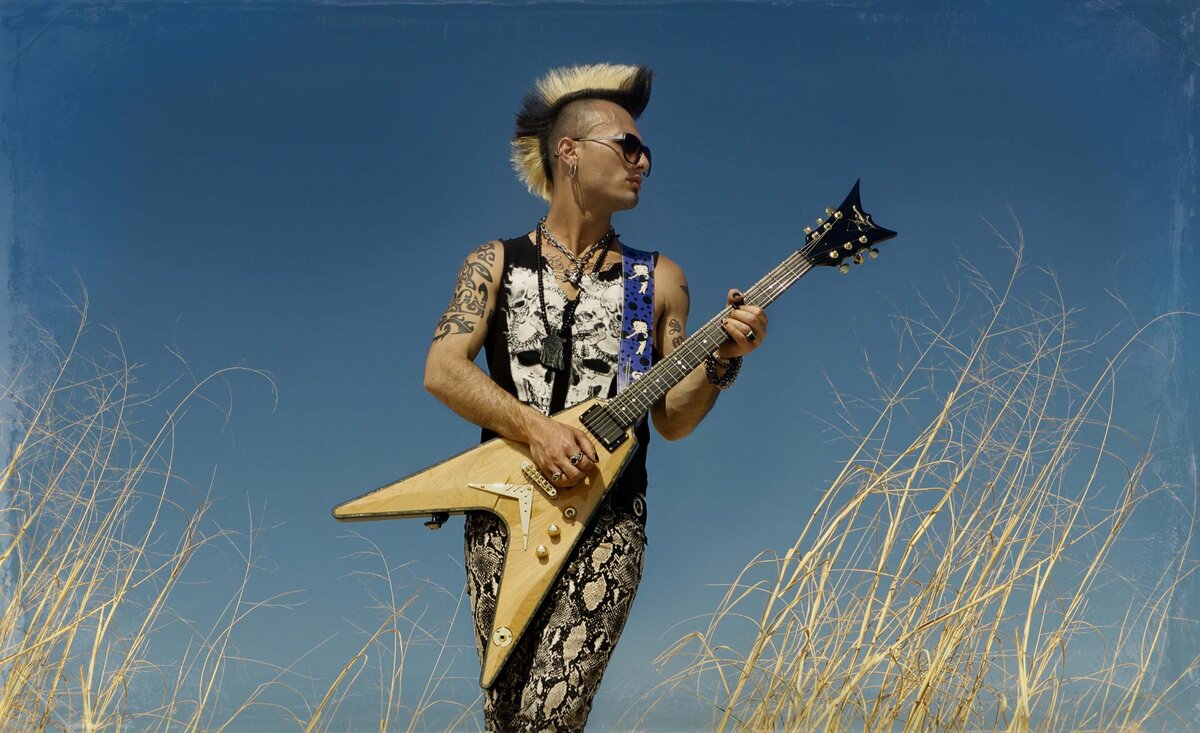 Male musician portrait Sal Costa wearing black and whites sleeveless pattern outfit holding gold electric guitar tall dry weeds beside