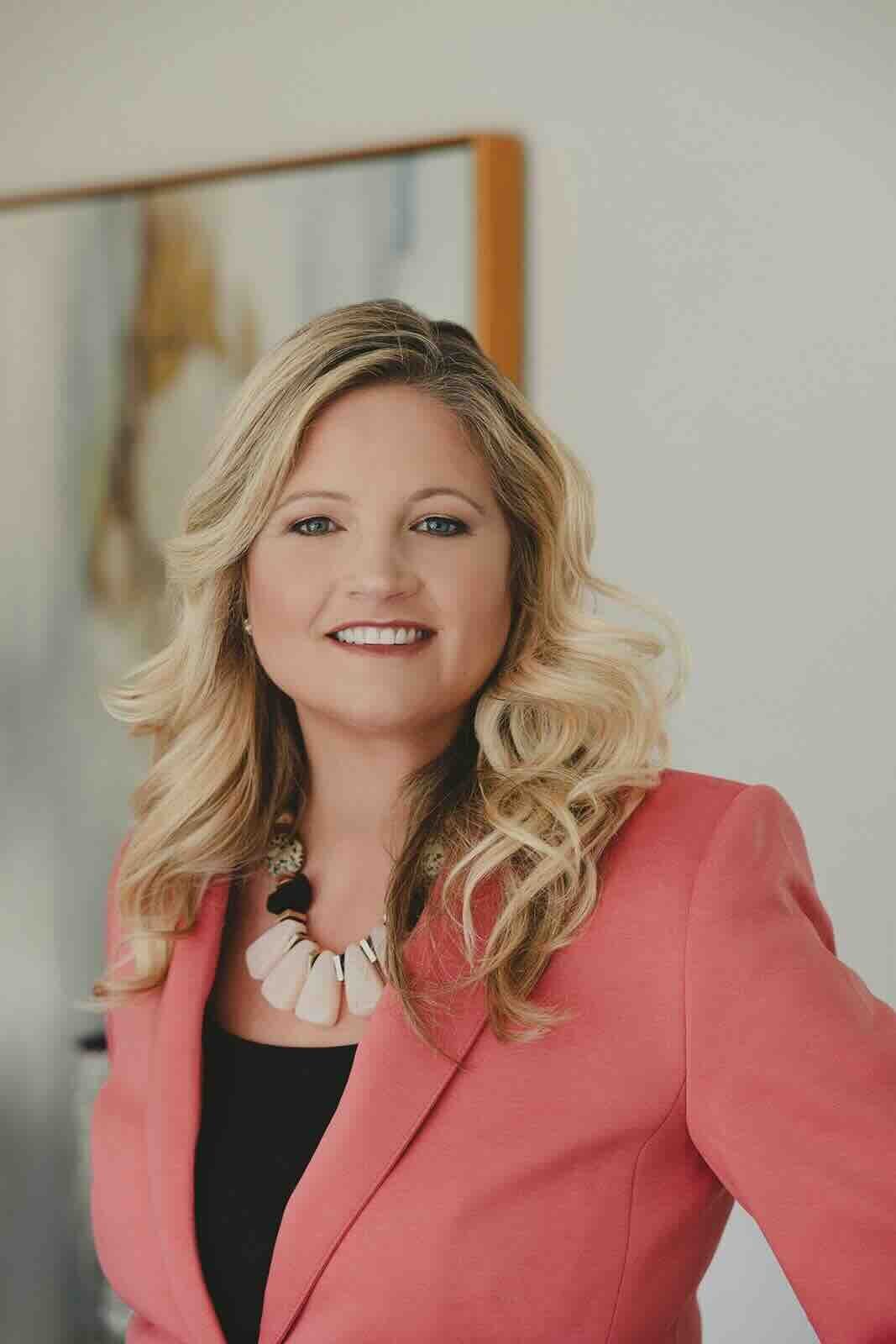 Professional branding photo of a woman in a coral blazer, smiling in a bright, welcoming office space.