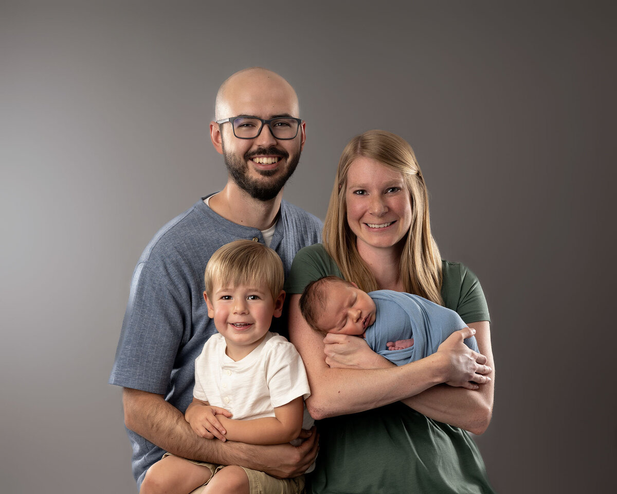 A happy family stands and smiles in the studio with a newborn and toddler boys