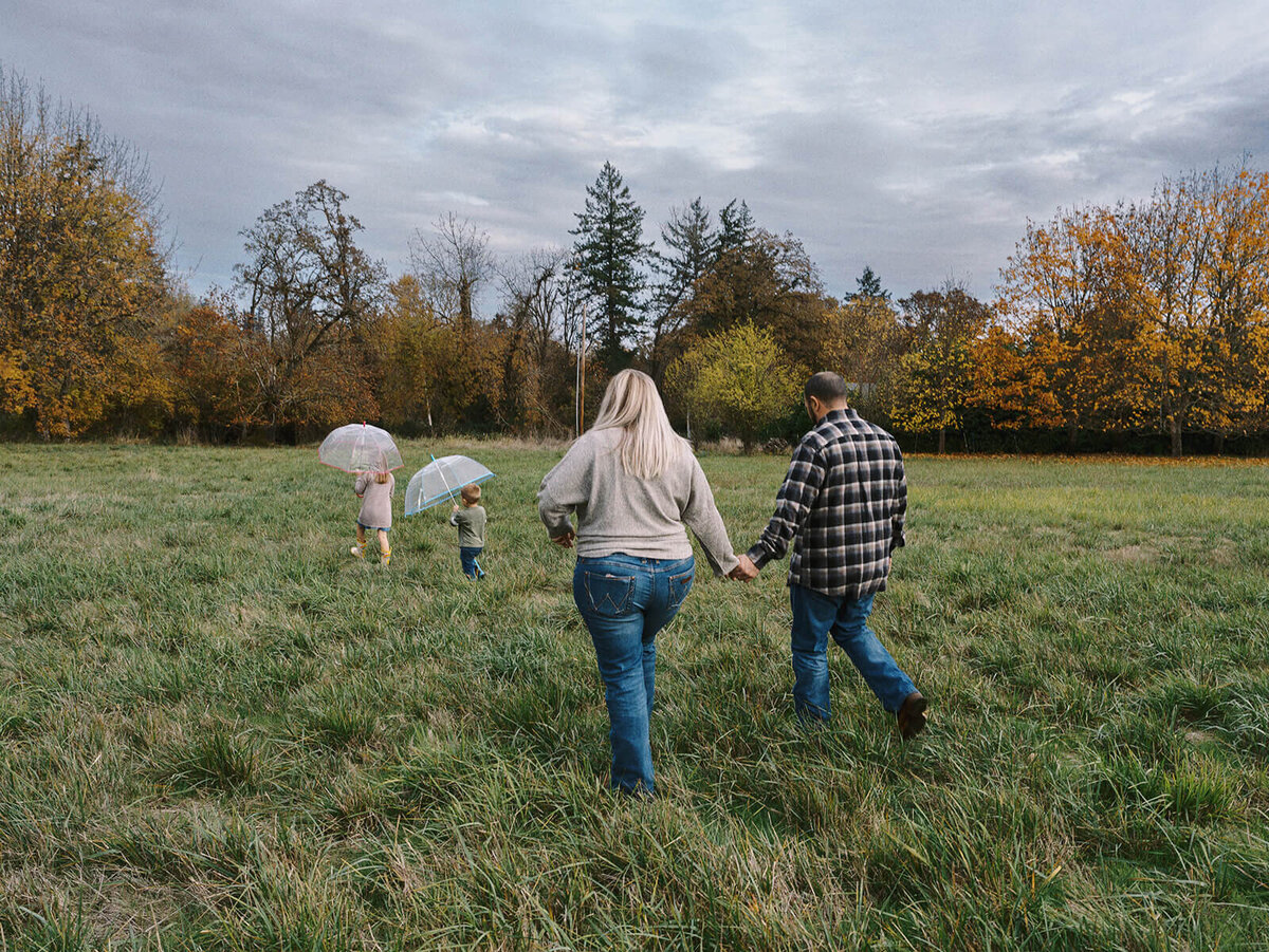 A family walks outdoors through a green field. The two kids run ahead of the parents holding umbrellas.