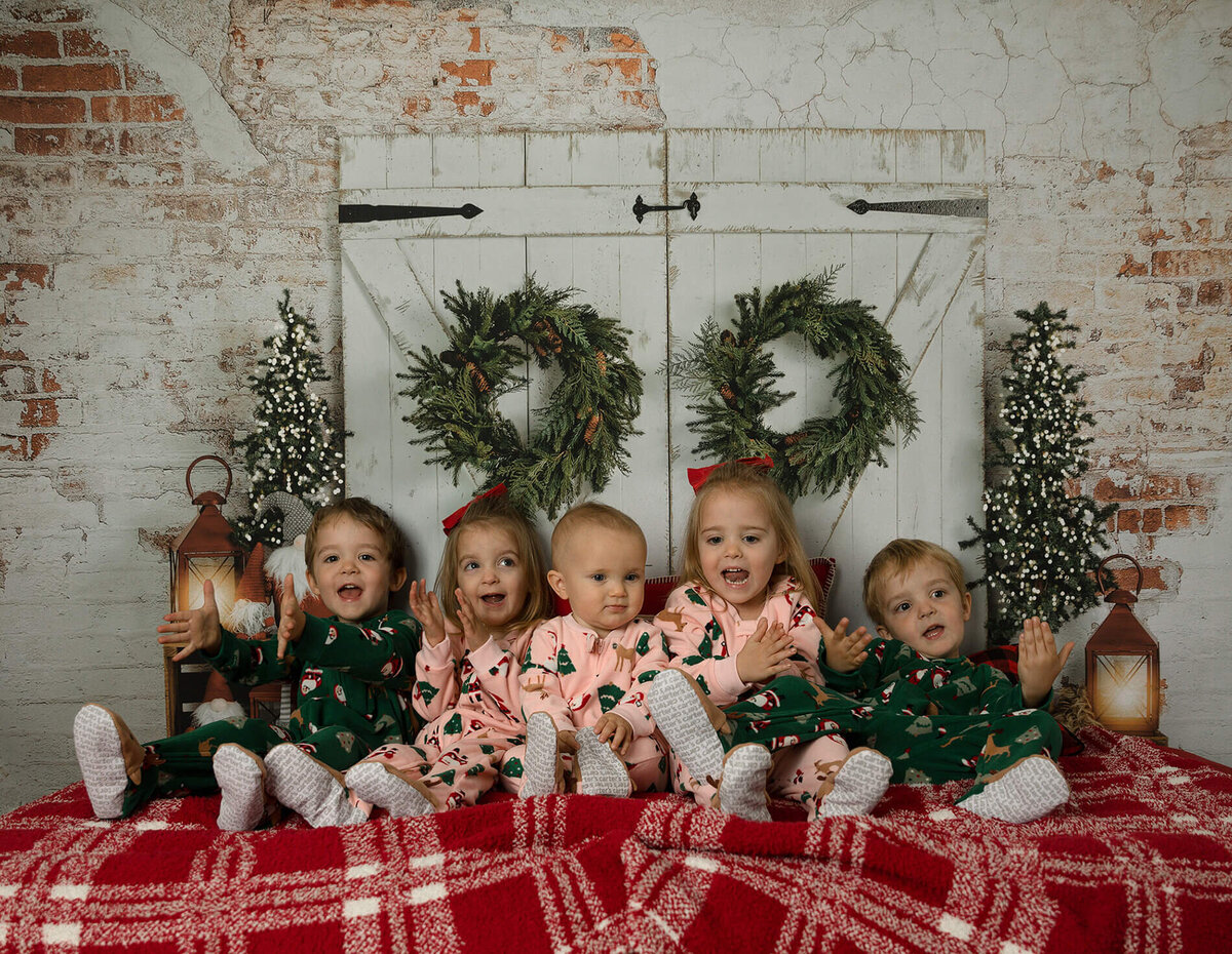 Quadruplets and their 1 year old sister all sitting on a bed in front of a Christmas background wearing their pajamas.