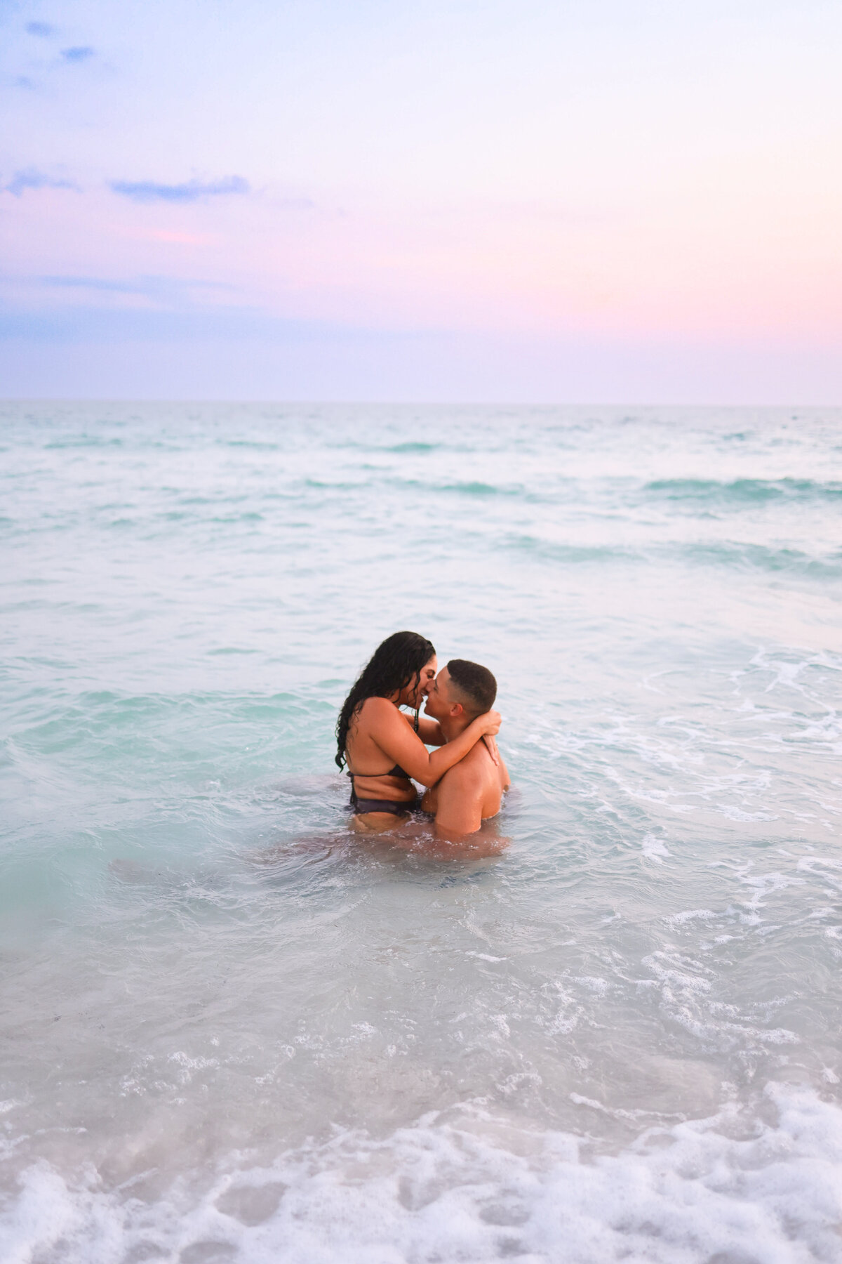 man and woman sitting in ocean kissing while wearing bathing suits