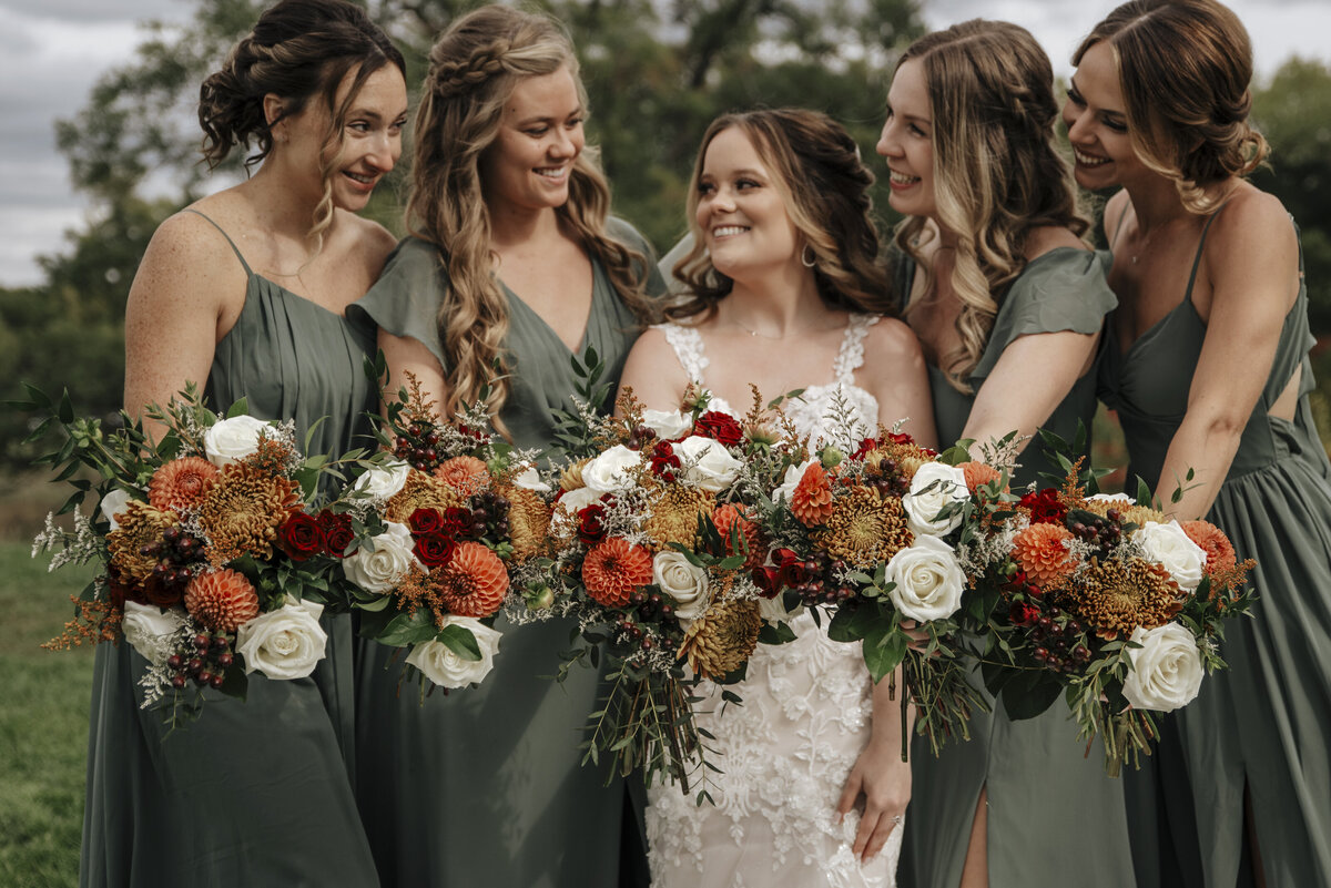 A joyful bride accompanied by her bridesmaids, all wearing coordinating green dresses and holding beautiful bouquets of flowers, sharing a moment of happiness together outdoors taken by jen jarmuzek photography, a minneapolis wedding photographer