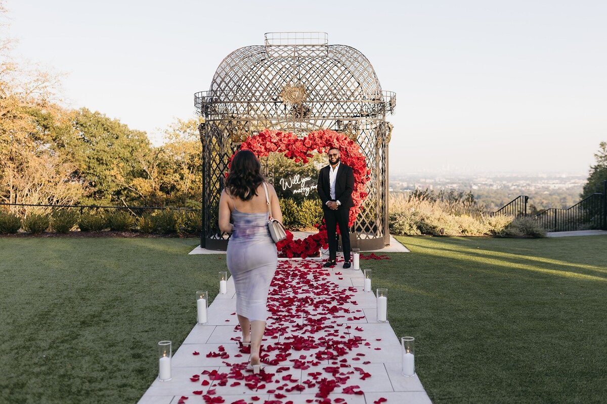 A romantic proposal setup designed by Yours Truly, a New Jersey wedding planner and designer, featuring a rose petal walkway, candlelit aisle, and a red heart-shaped floral arch.