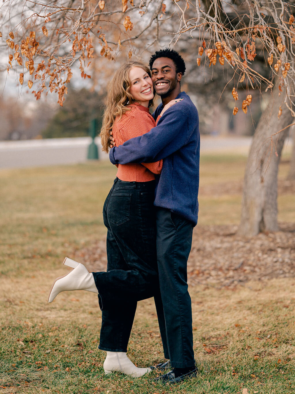 A couple stands under a tree at Cheesman Park. They are hugging each other, but looking at the camera, smiling.