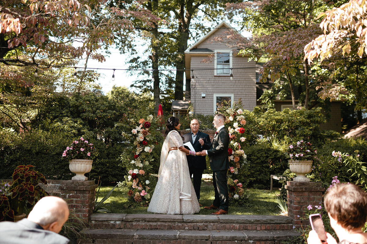 The couple standing before a floral arch, framed by a charming backyard garden and soft natural light.