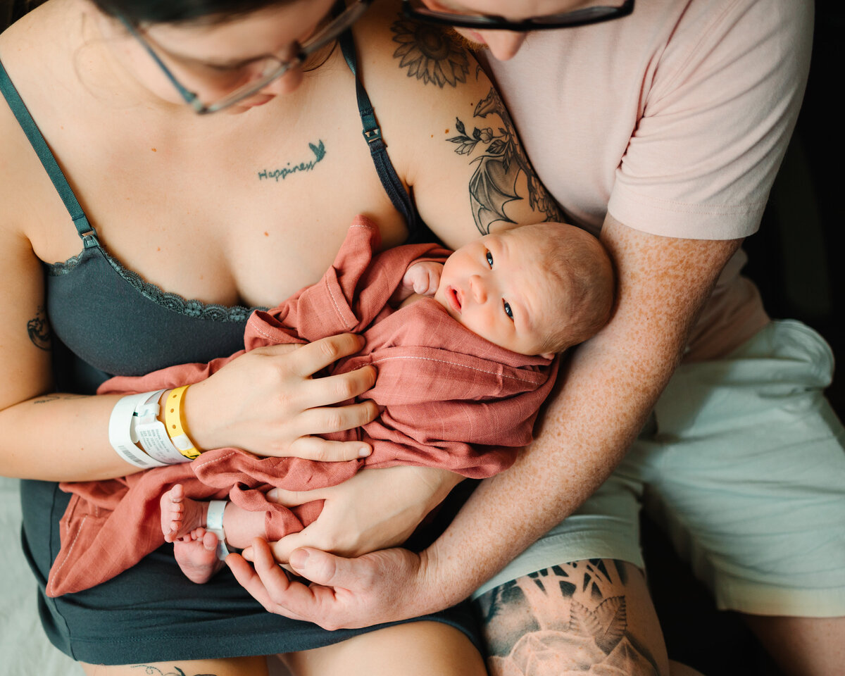 The love and joy of parenthood with Albuquerque maternity and newborn photography. This beautiful image shows happy parents admiring their newborn baby, who is wrapped in a pink blanket. The mother is dressed in a lovely green dress, while the father wears a pink t-shirt, reflecting the warmth and affection of the family