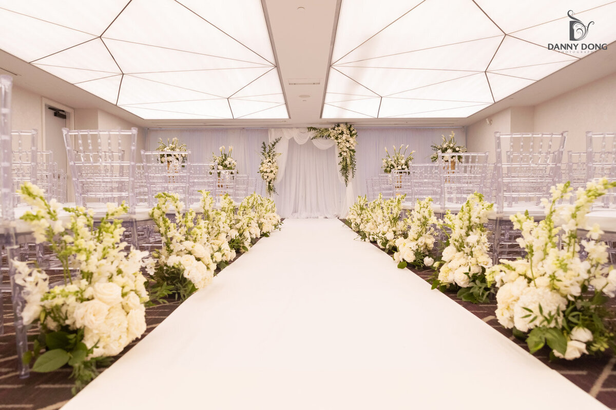 Indoor wedding aisle lined with white floral arrangements and draped backdrop