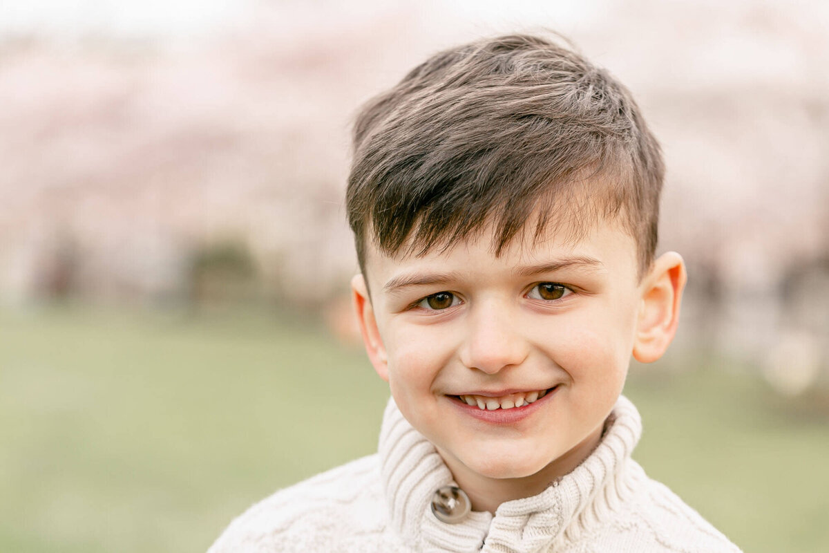 5 year old boy smiling at the camera with blooming cherry trees blurred out in the background behind him.