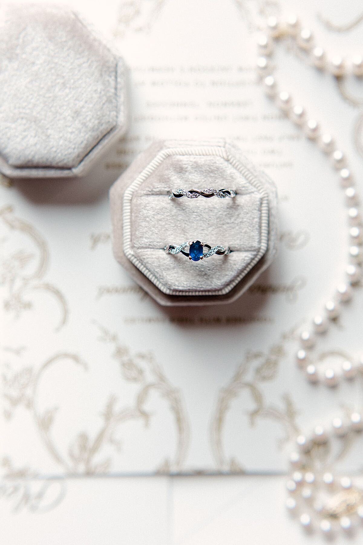 A white velvet ring box with an oval cut sapphire and diamond engagement ring and interwoven diamond wedding band on a table next to a pearl necklace.