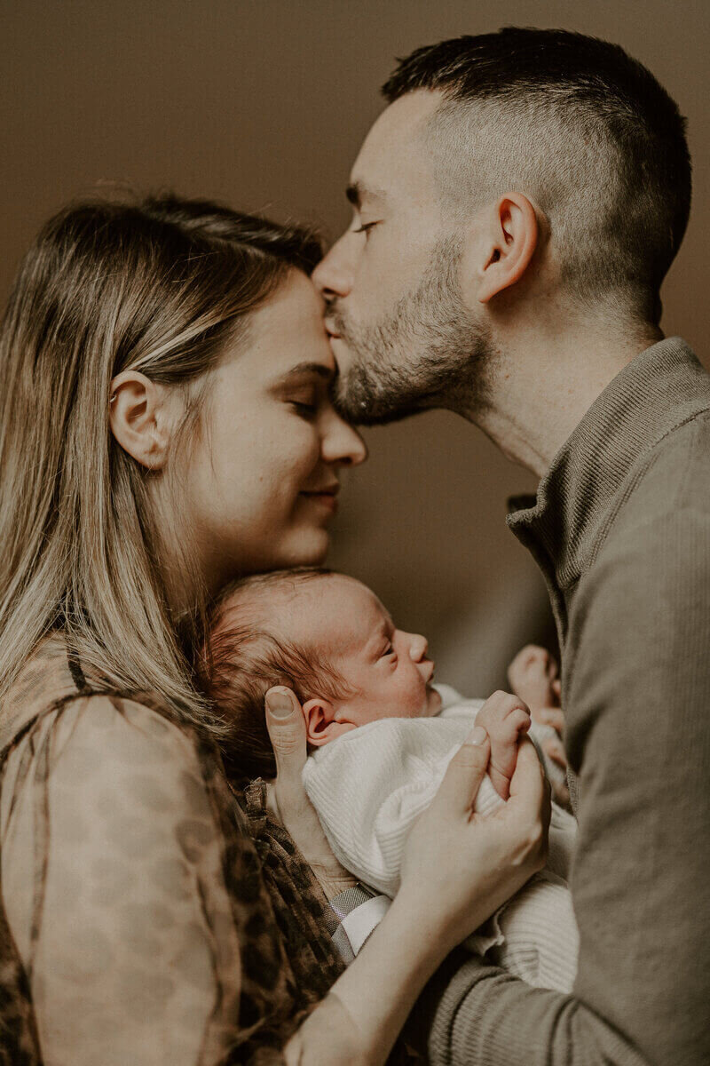 Homme embrassant le front de sa chéri. Entre eux leur bébé qu'ils portent dans leurs bras. Shooting photo naissance.