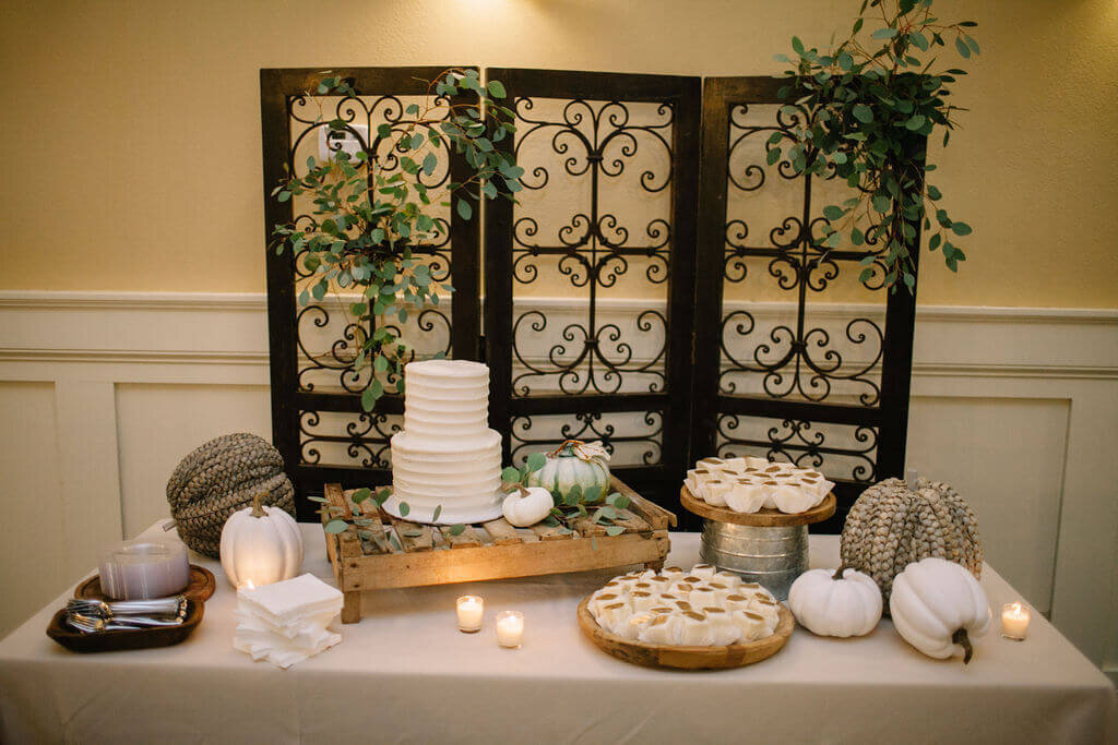 A rustic dessert table features a white tiered cake, mini pies, white pumpkins, candles, and greenery. The backdrop is a decorative wrought iron screen with leafy accents, creating an elegant and cozy setting.