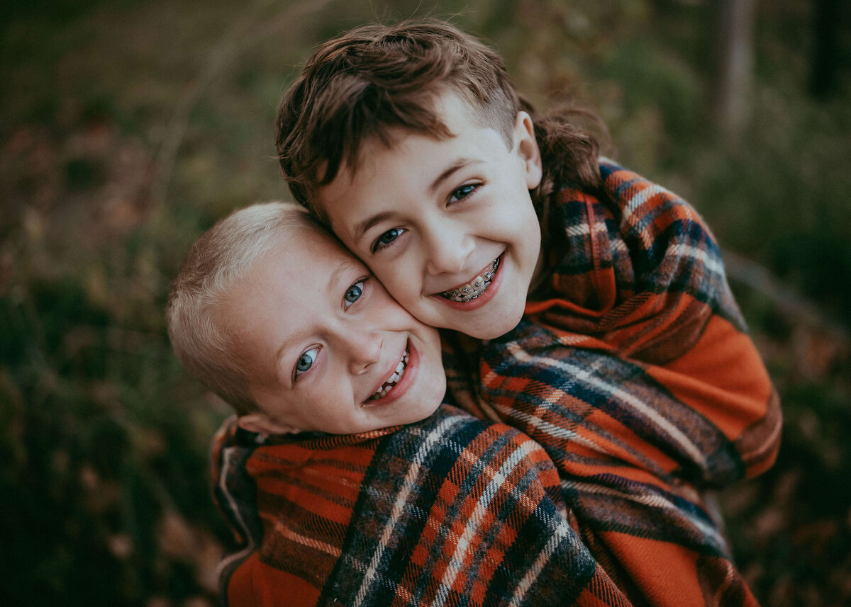 Two brothers squish their faces together as close as they can, while grinning at the camera being wrapped up like burritos in a plaid blanket on their family farm.