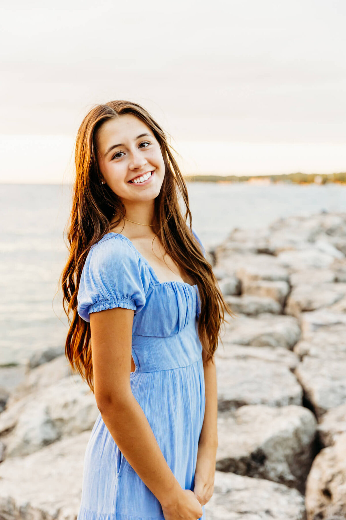 stunning high schooler in a light blue dress standing on a rocky shoreline  as the sun sets behind her in Sister Bay