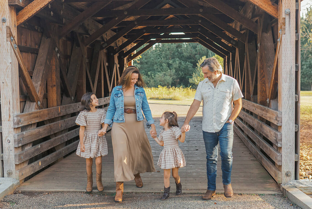 Family of four walking out of the covered bridge at Garrard Landing Park in Alpharetta, Georgia during sunset. Mom, dad, and their two young daughters smile and hold hands in the golden light, captured by Denay Shook Photography.