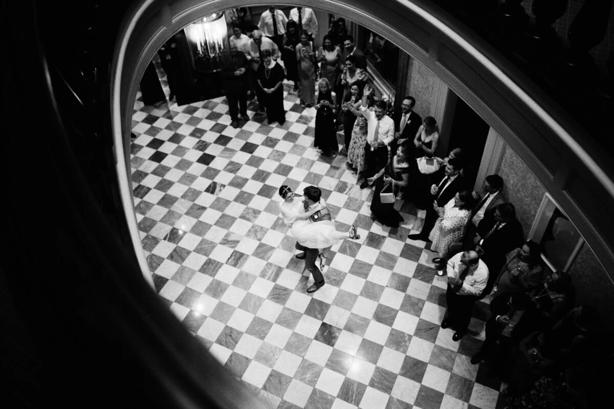 A couple dances in the center of a checkered floor, surrounded by a crowd in a grand room. Captured by a film photographer NJ, the black and white image, taken from above, exudes timeless elegance.