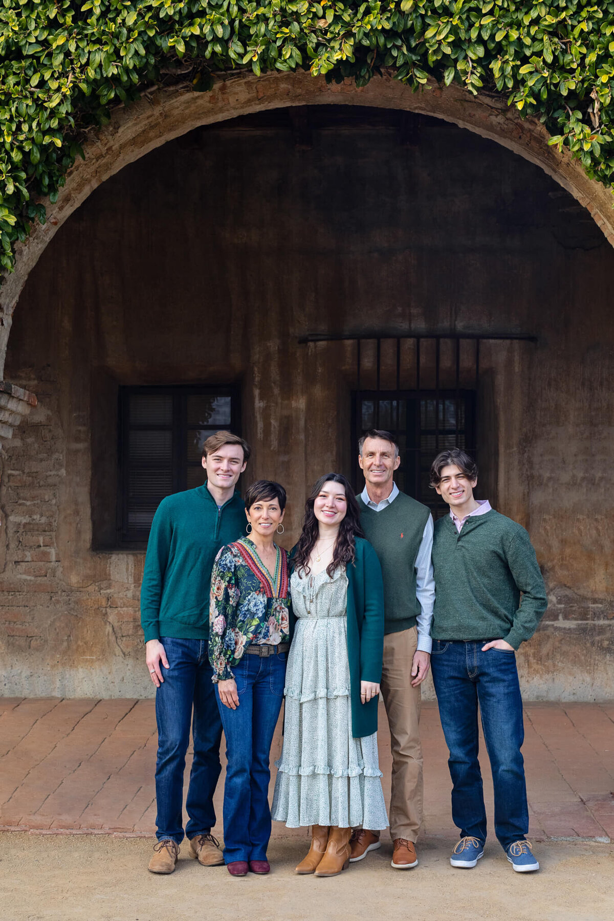 A family of five with dark hair wearing shades of green standing together underneath a brick archway with green vines on top of the arch. Captured at San Juan Capistrano.