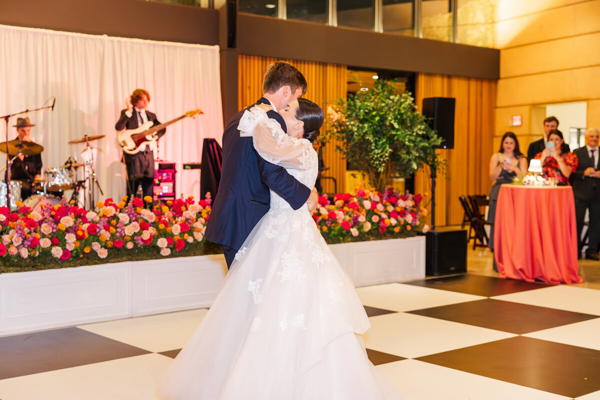Bride and groom embrace for their first dance while a live band plays behind them. The front of the stage is covered in beautiful pink flowers. The San Antonio wedding venue has a black and white checkered floor.