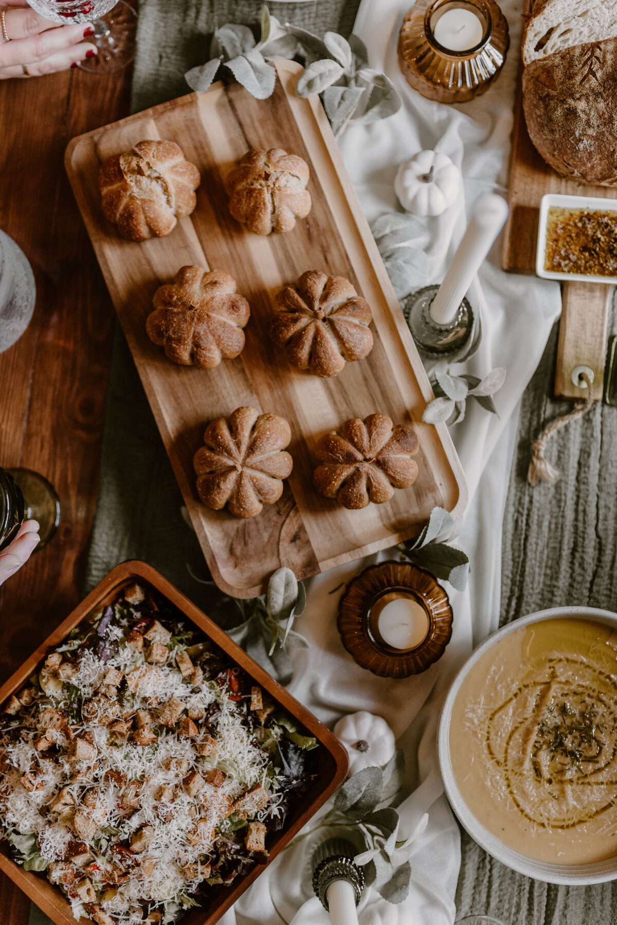 a table spread with delicious fall foods like soups, bread rolls, harvest salad, sourdough, and more. Dressed with candles and greenery, white and green table runner. 