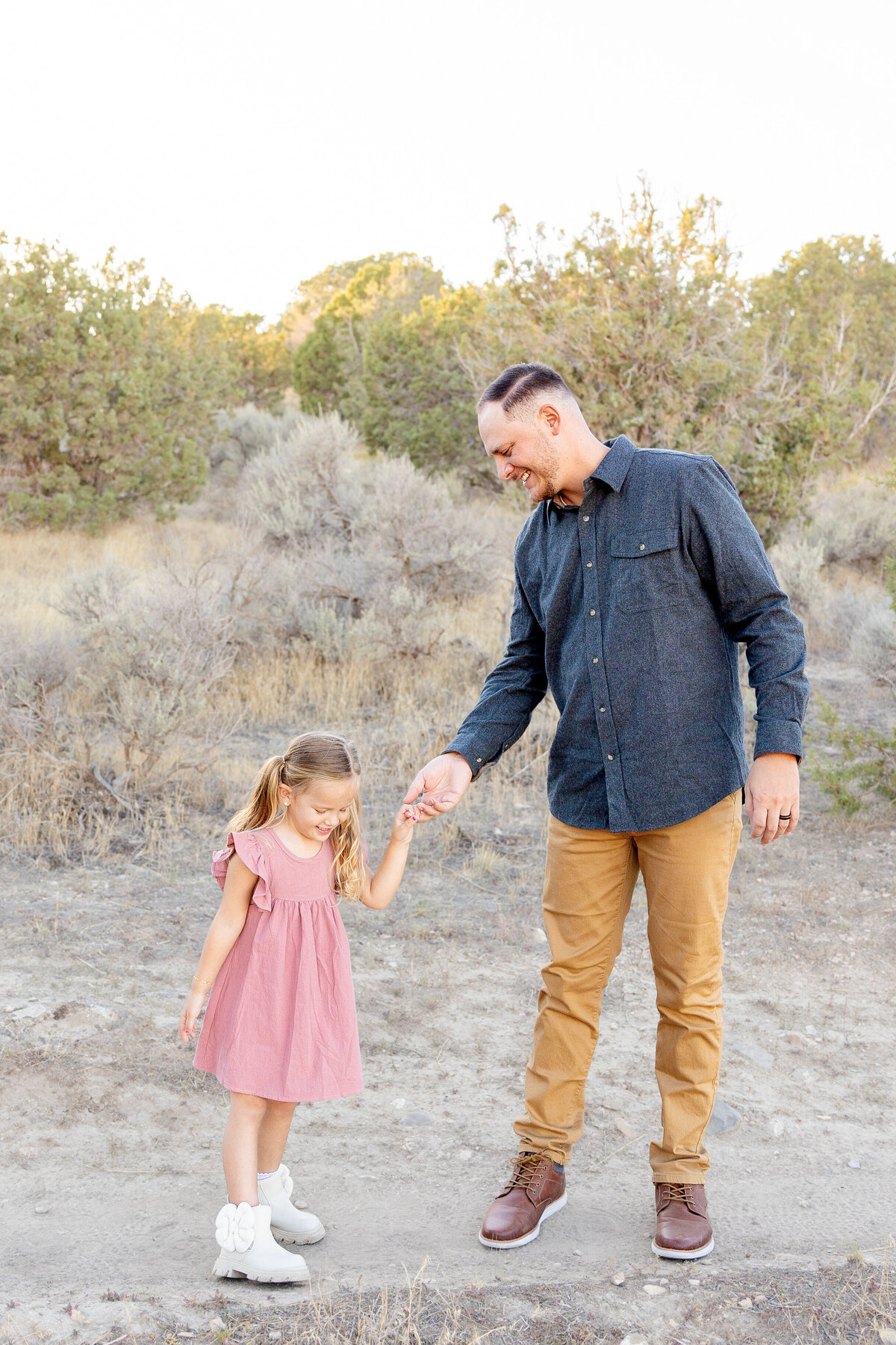 A father in a gray button-up shirt gently holds his young daughter’s hand as they walk together, sharing a happy and playful moment.