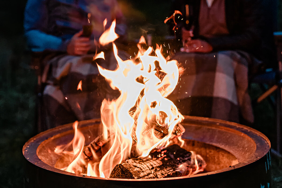A bride and groom sit behind a campfire, filled with crackling flaming logs in a Solo Stove during the evening portion of their two day luxury dogsledding and glamping elopement package  in Alaska.  They have a gray white and tan Pendleton blanket over their laps, and are holding a wine glass as they relax. 