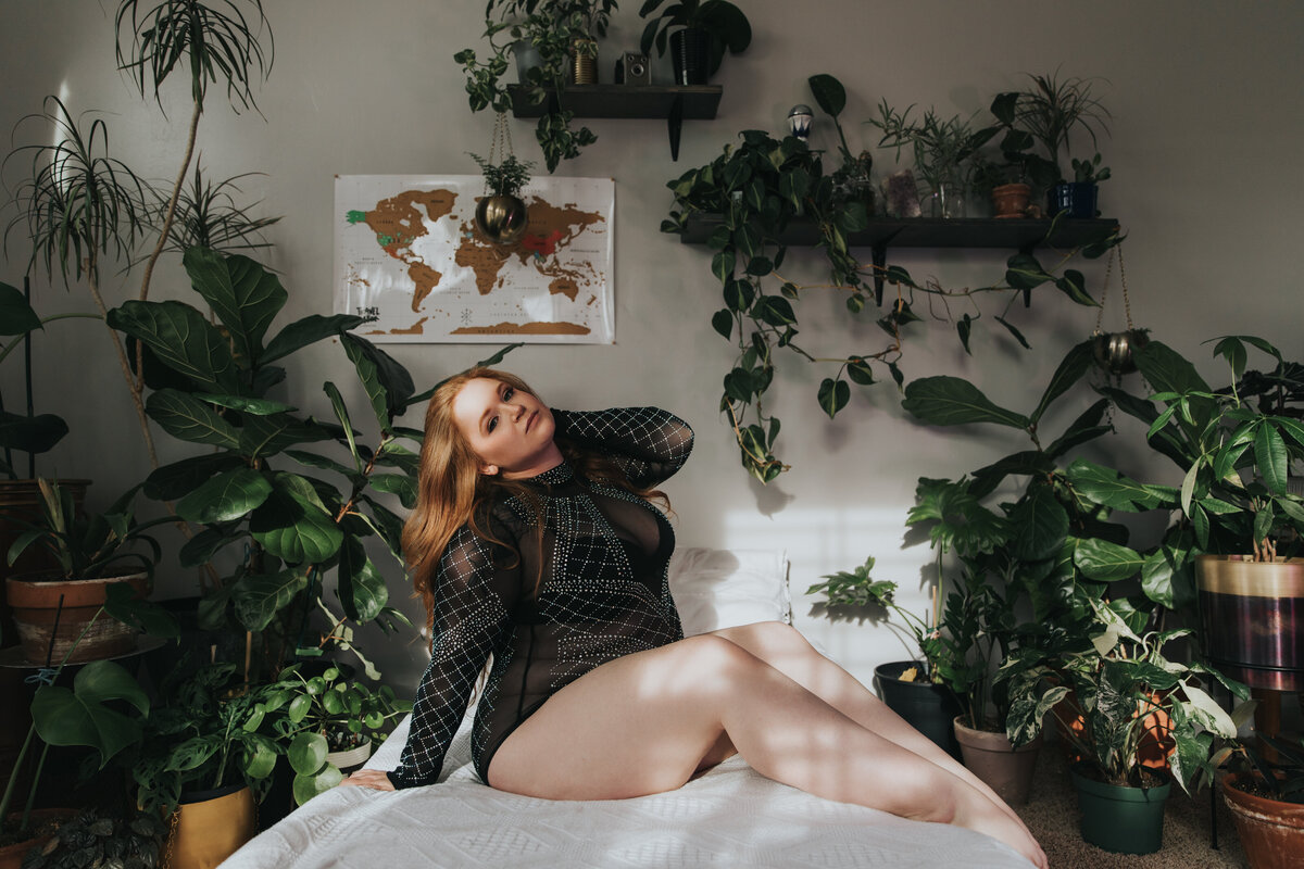 A red headed woman is sitting on a bed in the sunshine and she is surrounded by plants. Her legs are long and toes are pointed with one hand in her long hair