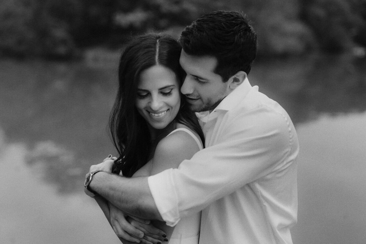 The man is lovingly hugging his fiancée from the back, at central park. A river is in the background. Image by Jenny Fu Studio