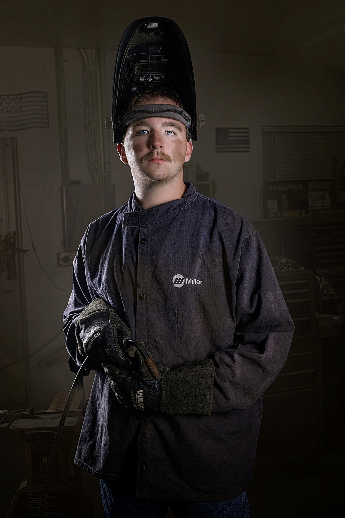 A high school senior stands in a welding shop in his jacket and helmet with his welder for his Pella Senior Photographer
