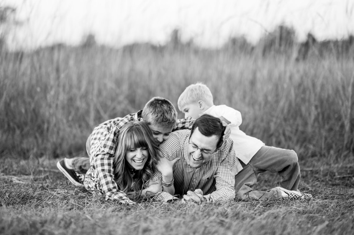 Husband and wife laying in a field with their two boys crawling over them.