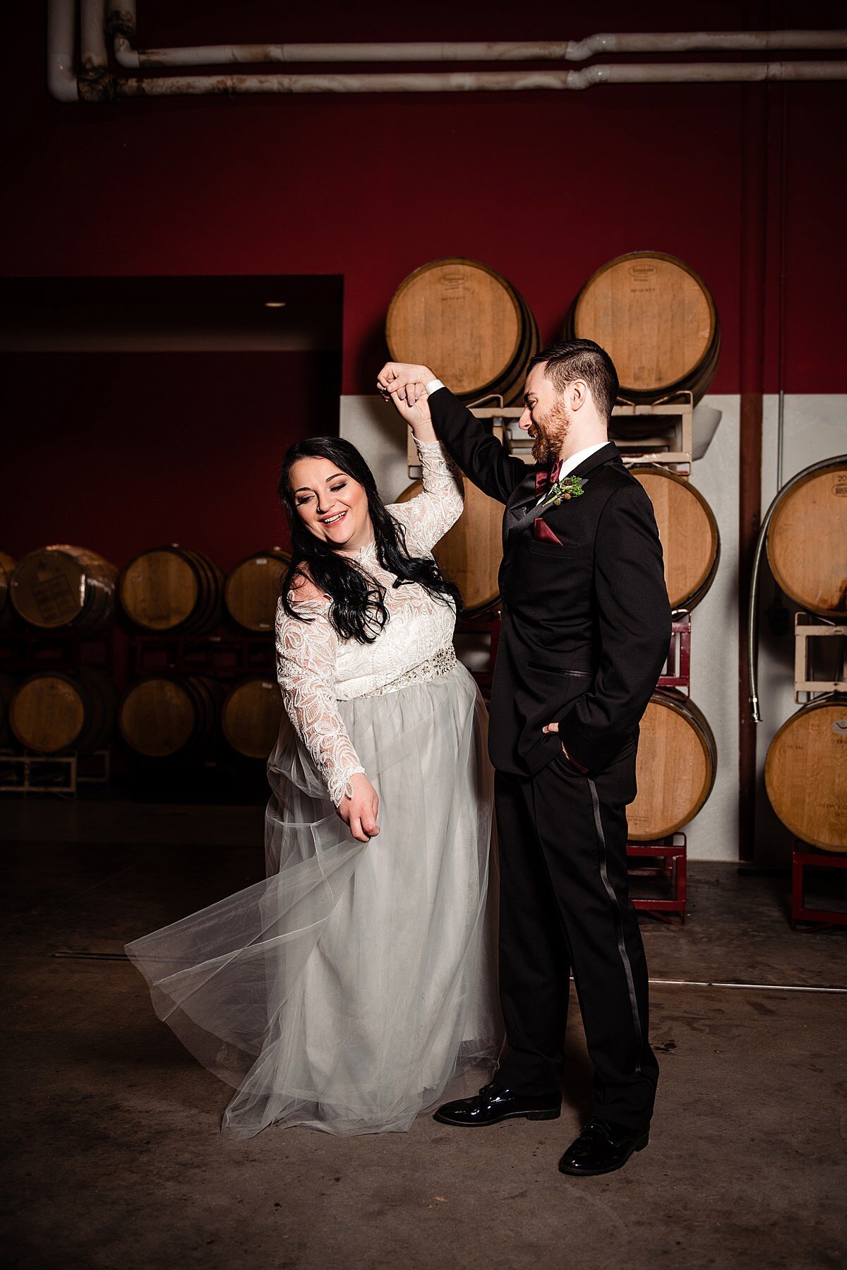 In the barrel room at City Winery, the groom, wearing a black tuxedo has one hand in his pocket  with the other hand raised above his head as the bride twirls in her wedding dress. The bride is wearing a fitted white lace top with a sheer grey tulle skirt. She is laughing as she spins.